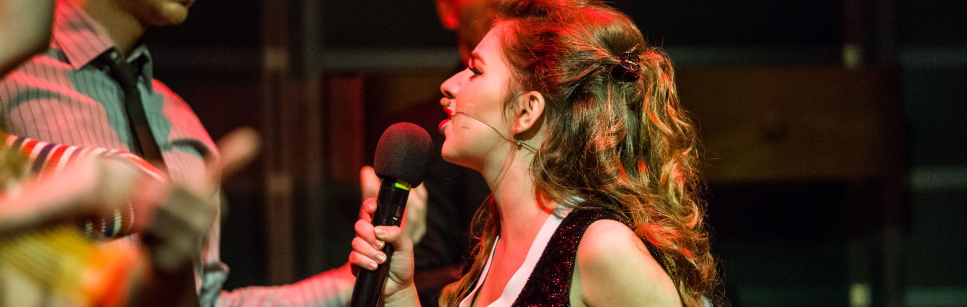 Młoda kobieta z profilu, trzyma w ręce mikrofon, wykłada jakby śpiewała do stojącego przed nią młodego mężczyzny w koszulki i czarnym krawacie.