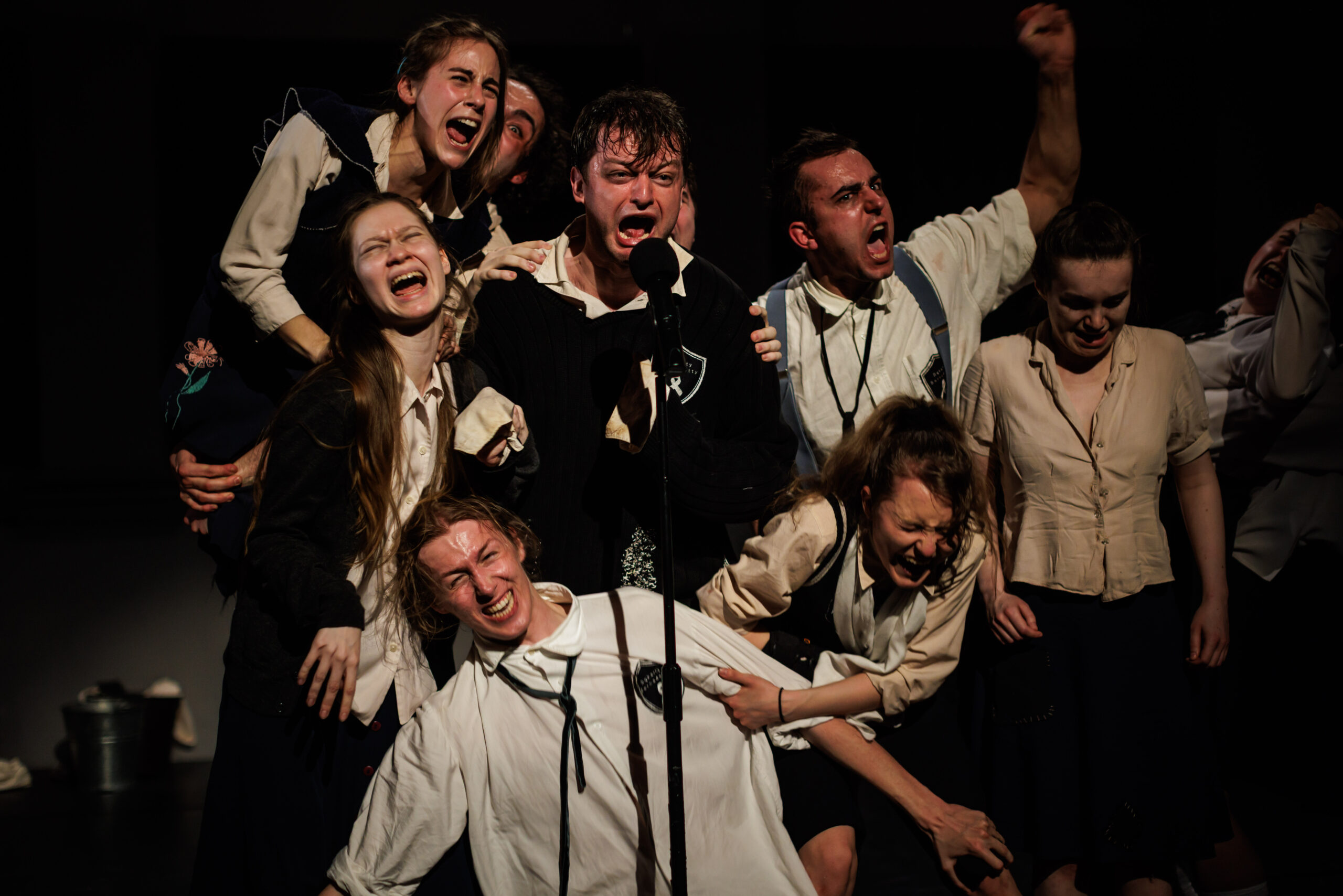 Grupa młodych ludzi w różnych konfiguracjach i pozycjach, starają się coś powiedzieć do mikrofonu. Przepychają się.