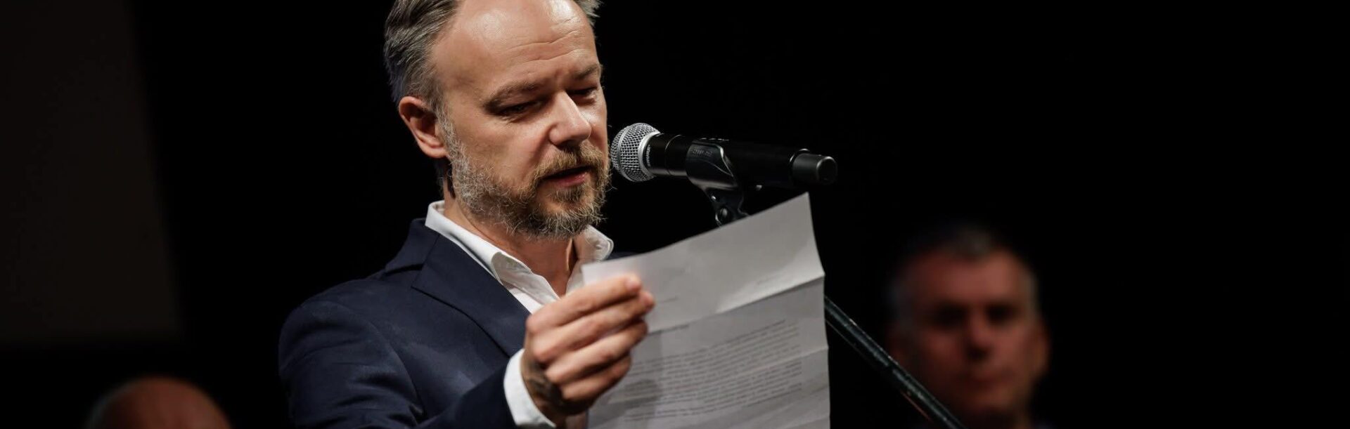 Dyrektor Szkoły Aktorskiej Marek Rachoń w garniturze, trzyma kartkę, przed nim stoi mikrofon.