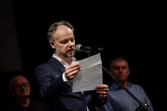 Dyrektor Szkoły Aktorskiej Marek Rachoń w garniturze, trzyma kartkę, przed nim stoi mikrofon.