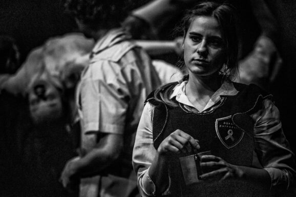 Kobieta na pierwszym planie miesza coś w metalowym kubku. Na drugim planie rozmyte ciała dzieci znęcających się nad swoją koleżanką.