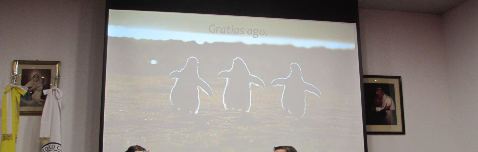 Michał Tadeusz Noworyta siedzi na stołem podczas konferencji, obok niego siedzi inny mężczyzna, za nimi na ekranie projektowa wyświetlają się trzy pingwiny oraz napis "Gratias ago".