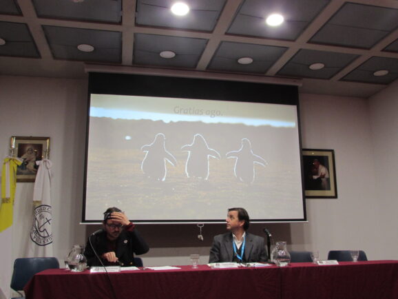 Michał Tadeusz Noworyta siedzi na stołem podczas konferencji, obok niego siedzi inny mężczyzna, za nimi na ekranie projektowa wyświetlają się trzy pingwiny oraz napis "Gratias ago".