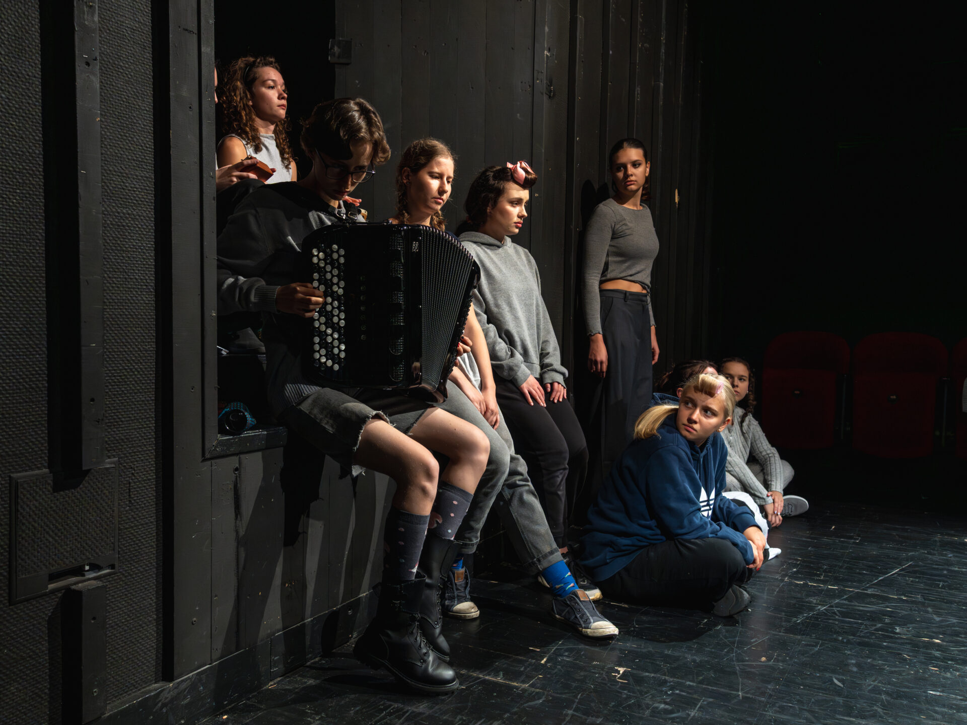Kilka osób, niektórzy siedzą na ziemi, niektórzy we wnęce Sceny Kameralnej Teatru Śląskiego, jeden chłopak trzyma na kolanach akordeon i gra na nim.