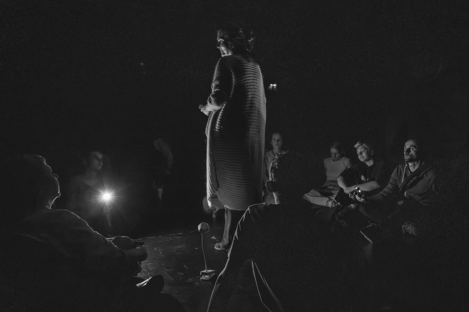 Kobieta stoi na środku odwrócona plecami. Wokół niej po turecku, w kole, siedzą inne osoby.