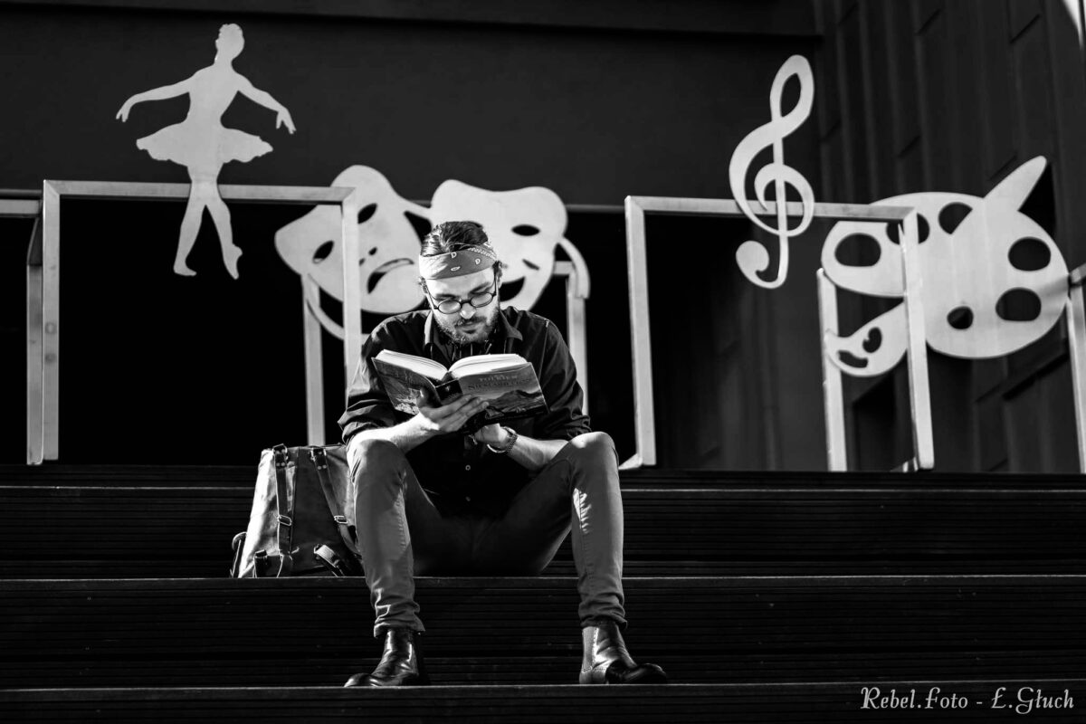 Portret Michała Tadeusza Noworyty. Siedzi na schodach z książką w ręku, za nim scenografia składająca się z symbolów: baletnicy, maski teatralnej, klucz wiolinowego oraz akcesoriów malarskich.